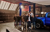 The cast for the promotion standing next to a blue raptor in a garage