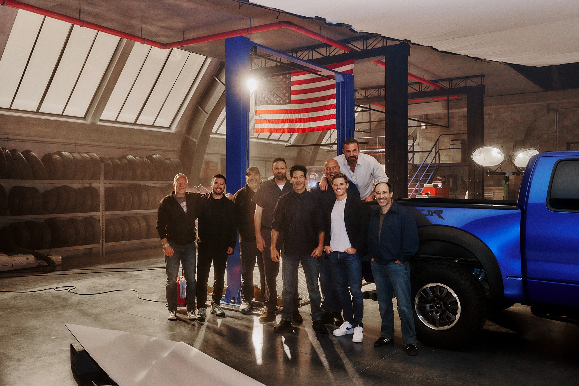 The cast for the promotion standing next to a blue raptor in a garage