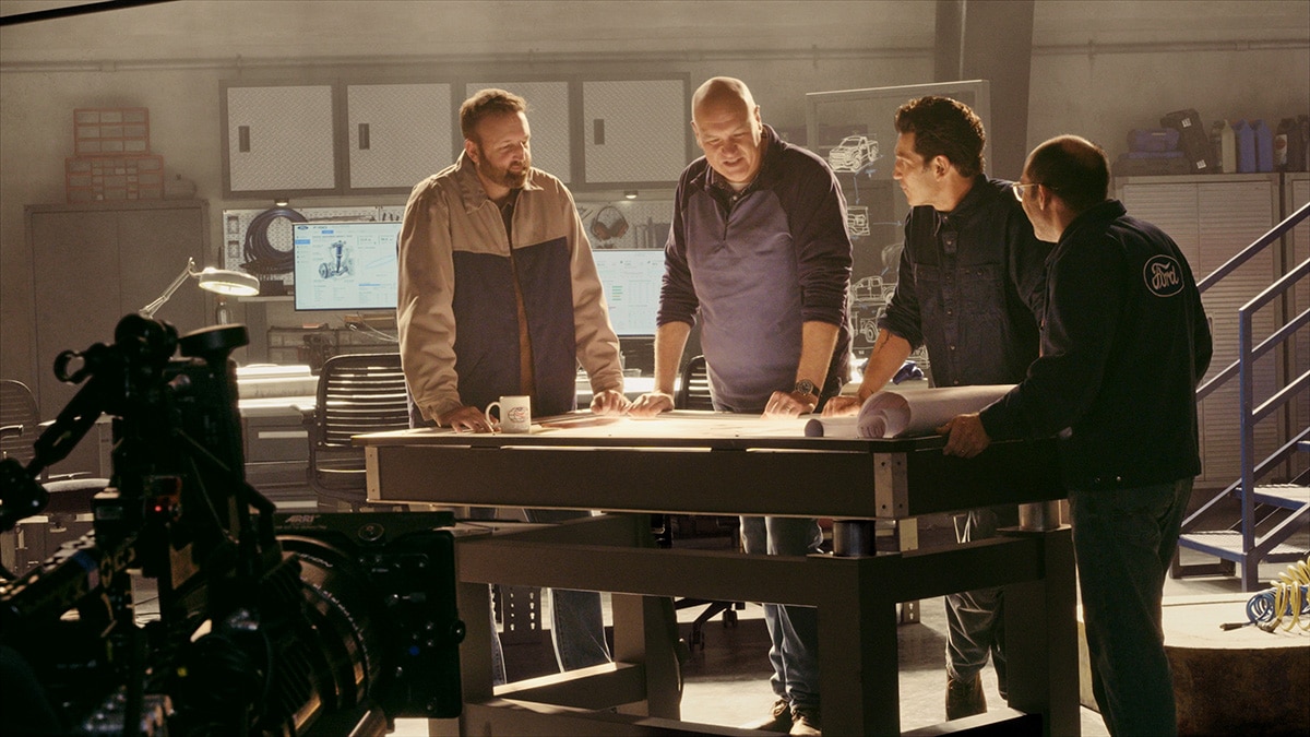 Wide angle of Jon Bernthal and the Ford Racing engineers standing over plans at a table