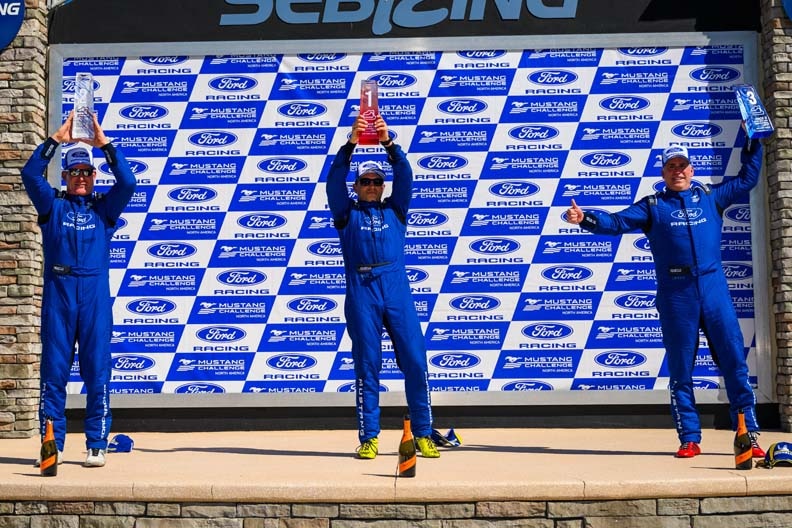 Three race car drivers stand on a winner's podium, holding trophies overhead