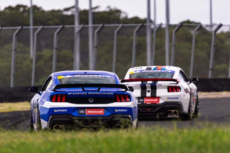 Three race car drivers stand on a winner's podium, holding trophies overhead