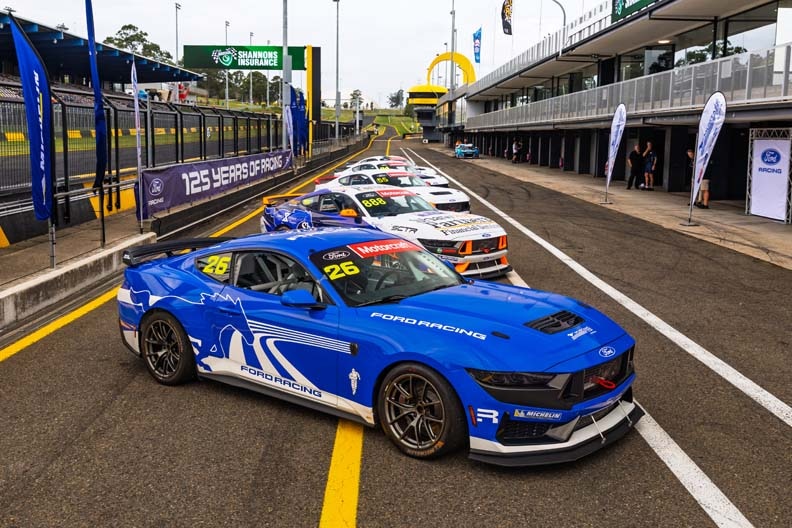 White, red, and black race car leading other race cars on a curved track with hills in the background