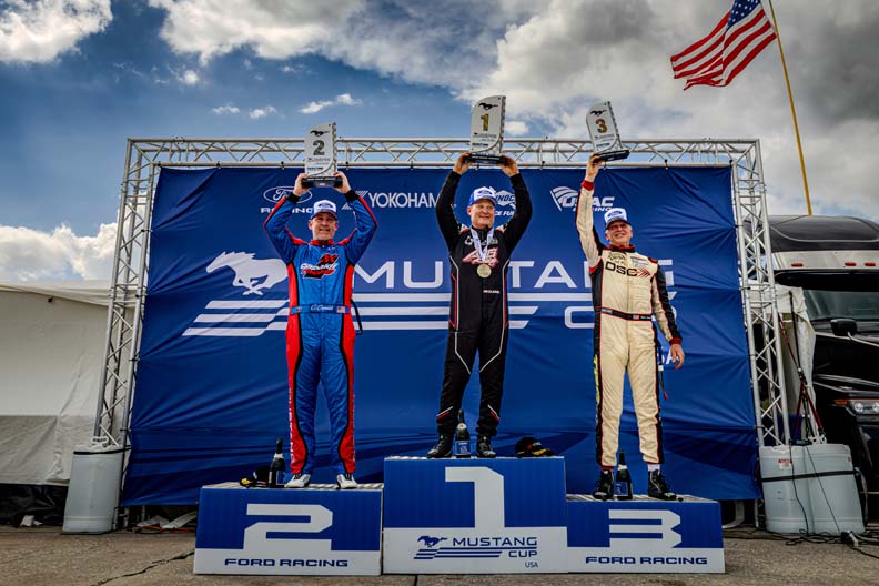 Three race car drivers stand on a winner's podium, holding trophies overhead