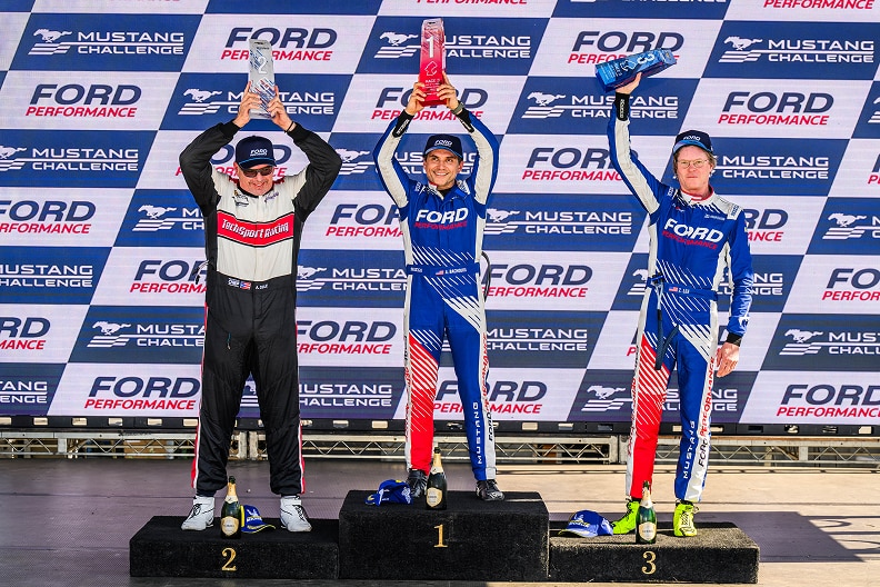 Three race car drivers stand on a winner's podium, holding trophies overhead