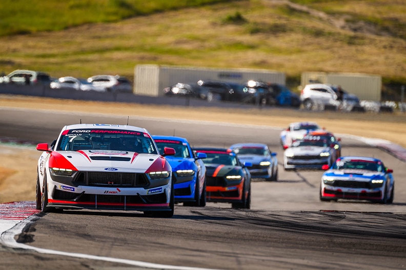 White, red, and black race car leading other race cars on a curved track with hills in the background