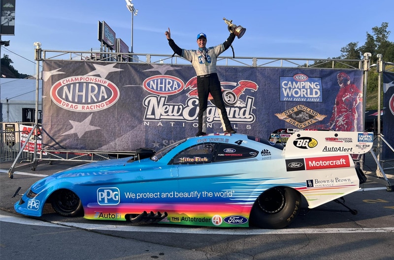 Bob Tasca III standing on top of NHRA Drag Car with trophy