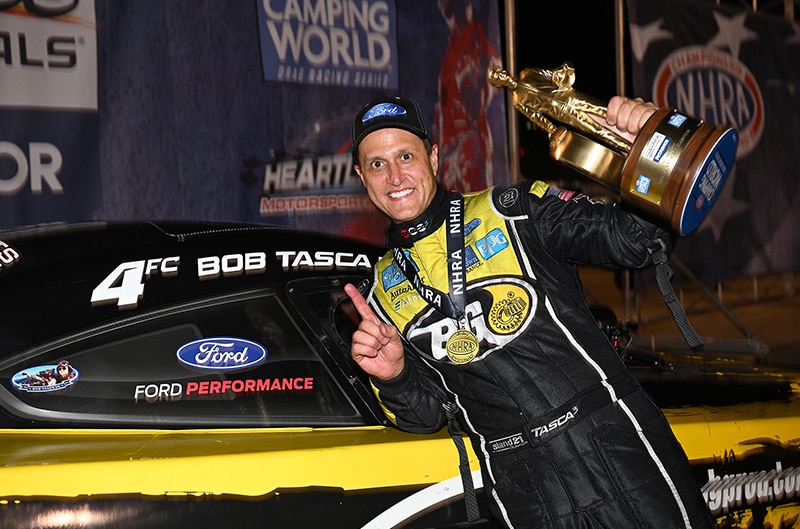 Bob Tasca III with trophy posing next to Ford Performance logo on drag car